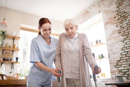 A caregiver in scrubs assists an elderly woman using crutches in a well-lit kitchen with shelves and brick walls in the background.