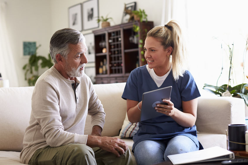 A healthcare professional in blue scrubs sits on a couch with an older man, showing him a tablet. They are engaged in conversation in a living room setting.