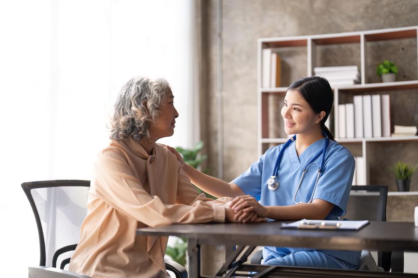 A healthcare professional in blue scrubs holds the hand of an elderly person with gray hair, providing comfort and support at a table in a well-lit room with a bookshelf in the background.