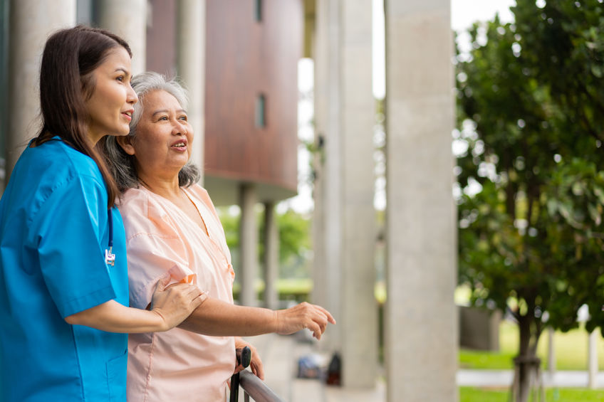 A younger woman in a blue uniform stands beside an older woman in a peach blouse, both looking outwards on a balcony with large columns and greenery in the background.