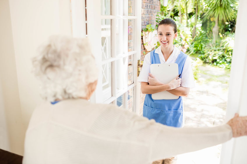 A nurse holding a clipboard stands at the door of an elderly woman’s home, smiling as she is welcomed inside.