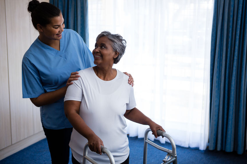 A caregiver in a blue uniform assists an elderly woman using a walker. They are indoors near a large window with blue curtains.